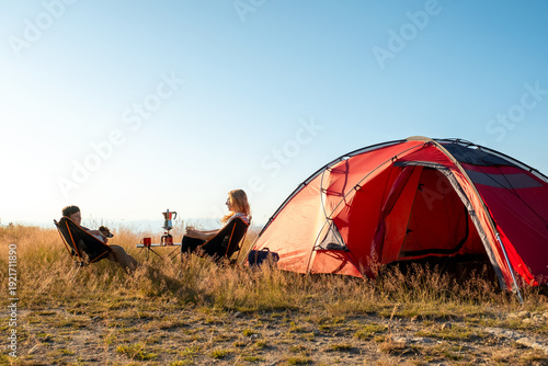Morning evening ritual camping mountains mother son talking sitting chairs near red tent. Scenic wilderness background sun rays. Pet dog companion. Authentic travel experience family togetherness natu