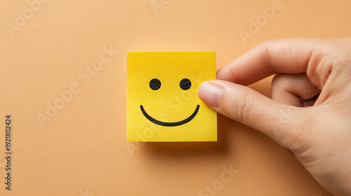 A hand holds a yellow sticky note with a simple black smiley face against a beige background.