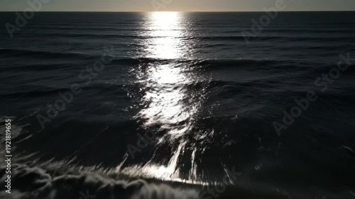 Ocean waves gently rolling towards the shore under a bright sun reflecting on the water surface, creating a serene coastal atmosphere at dusk