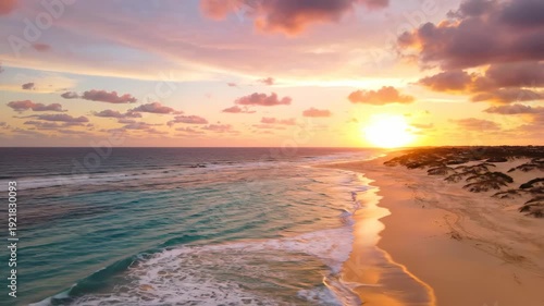 Sunset over tranquil beach with gentle waves lapping at the shore, colorful clouds reflecting warm hues in the sky, and sand dunes visible in the background