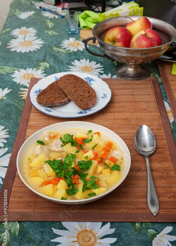 Vegetables and Meat Soup with Pork and Cooking Roots: Potato, Carrot, Celery, Turnip and Parsley and Rye Bread in Plates on Mat and Apples in Metal Vase on Kitchen Table. 