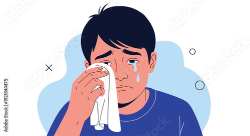 Sad young man with dark hair cries and wipes tears from his eyes with a white tissue while feeling emotional and distressed on a blue background.