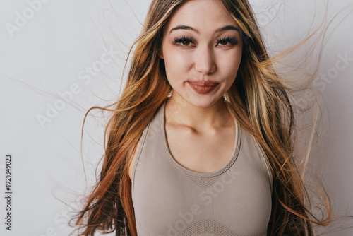 Young woman standing with long hair blowing in the wind wearing sleeveless top looking confidently at camera smiling softly indoors with neutral background.