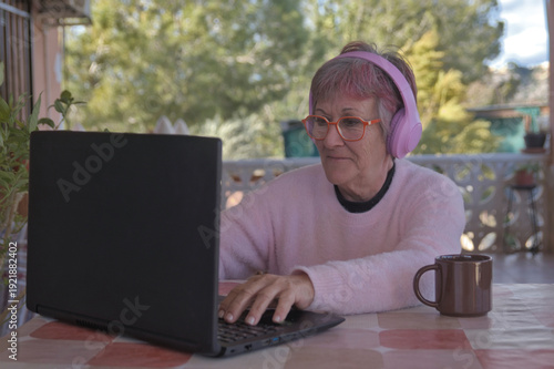 Older woman with headphones happily using her laptop in her country house