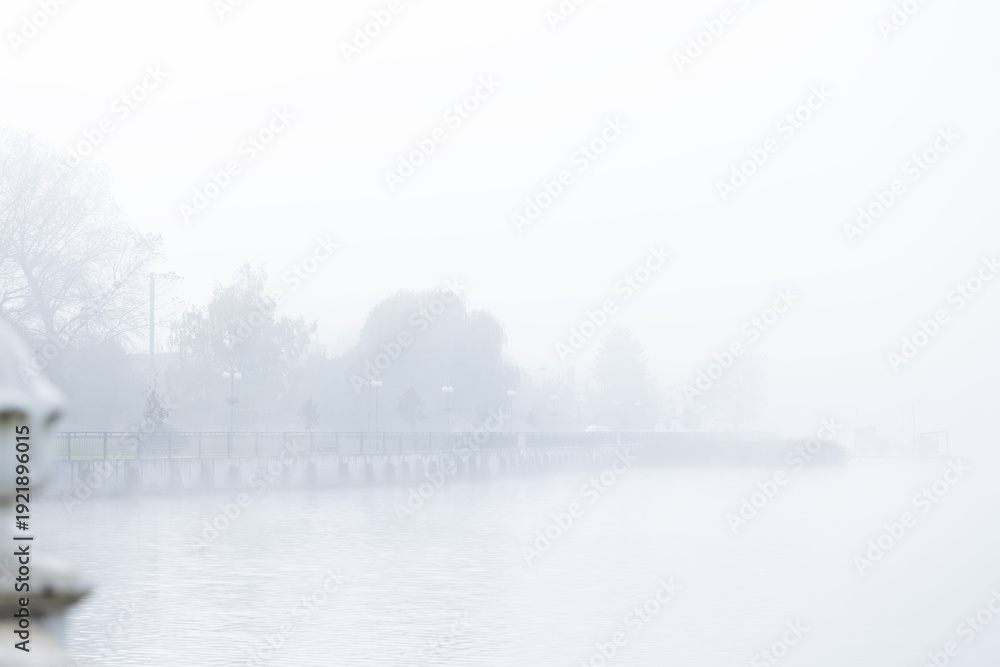 Fototapeta premium Misty morning over a calm lake with a distant pier and trees shrouded in fog