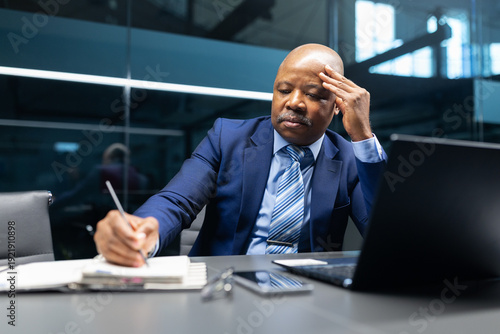 Φωτογραφία Mature black businessman writing notes while reviewing information on laptop in modern office