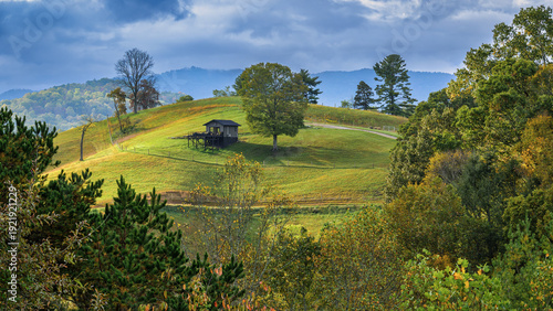 A small, dark wooden cabin with a deck sits on a grassy hillside under a cloudy sky. The surrounding landscape is dotted with trees, some showing the colors of autumn, and rolling hills recede into