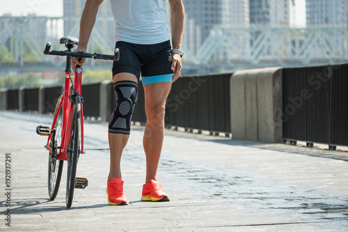Fotografie Close up of male cyclist leg with knee brace walking with bicycle