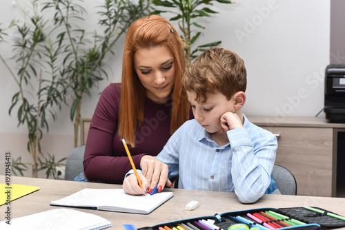 Caring mother assisting her little boy with school homework. Child studying with parent at home using pencils and notebook. Education, childhood development, homeschooling and family bonding concept.