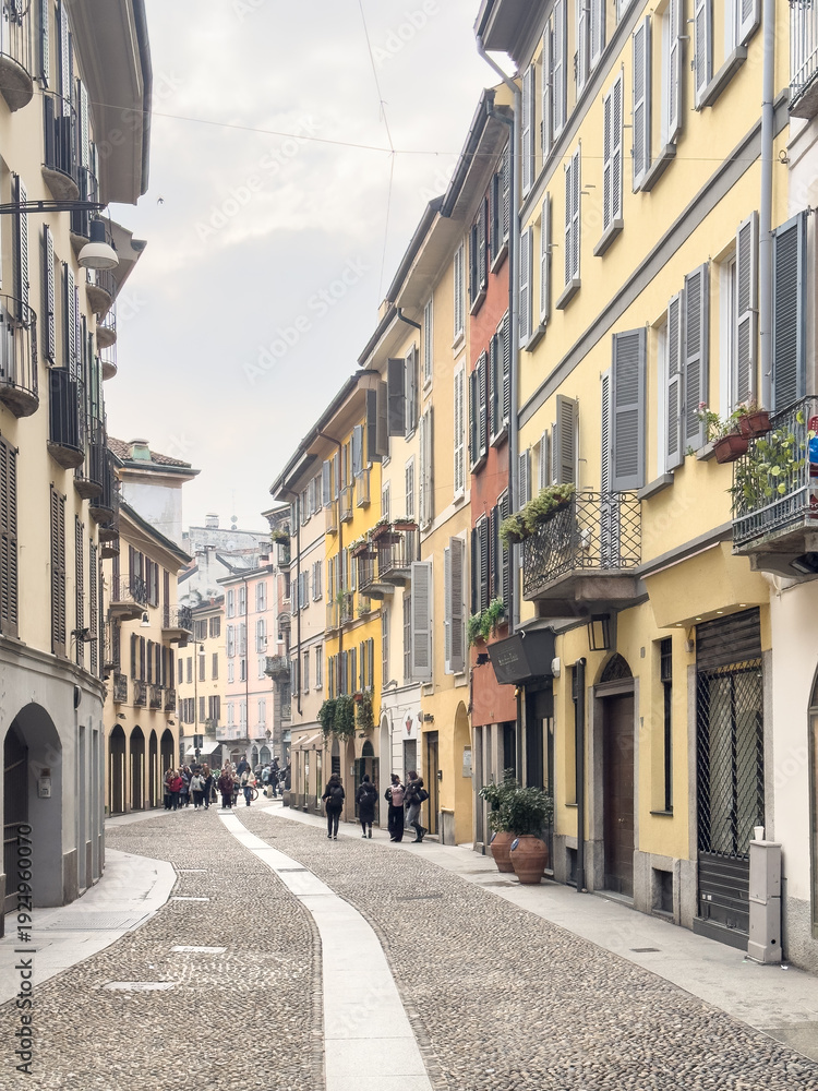 Obraz premium Colorful pastel buildings with shuttered windows line a curved cobblestone street in Milan, Italy with pedestrians in a historic neighborhood.