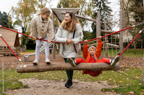 Mother guiding kids balancing on wooden playground rope bridge swing