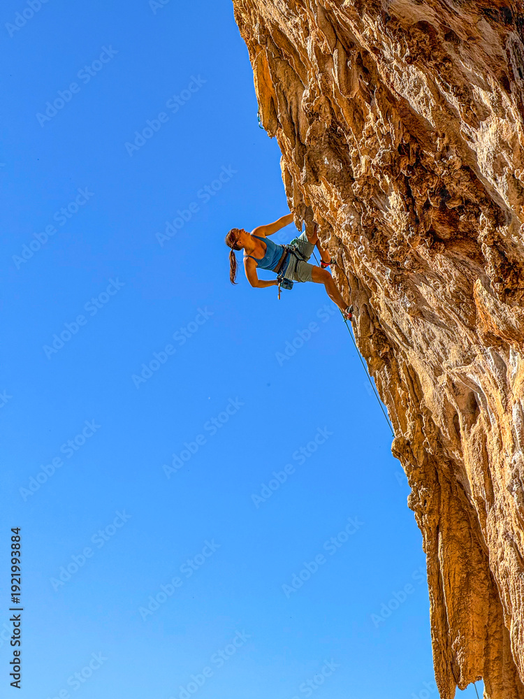Fototapeta premium LOW ANGLE VIEW, CLOSE UP: Strong female climber on steep limestone wall with tufa formations. Young woman reaches into her chalk bag while lead climbing a steep overhang under blue Mediterranean sky.