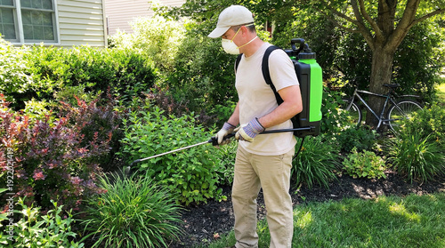 Pest control professional spraying pesticides on garden plants with backpack sprayer, wearing protective gear, outdoor residential setting