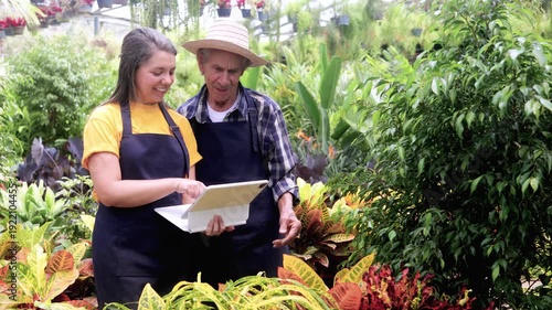 Hispanic multigenerational team using tablet at greenhouse during spring - Latin American woman teaching senior farmer