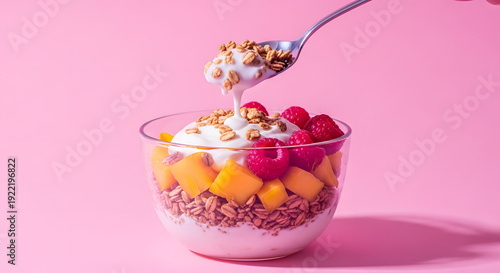Spoon scooping yogurt and granola from a healthy breakfast parfait. Fresh fruit bowl with mango and raspberry on a pink background with copy space