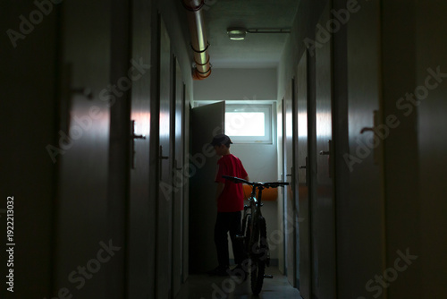 Принти на полотні teen boy with bicycle in corridor, red jacket catching fluorescent light, hands on handlebars while pausing before exit, quiet urban return from school or afterschool ride