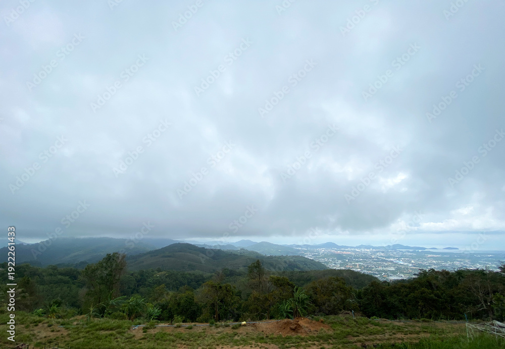 Obraz premium Scenic view of lush green hills and distant coastline under cloudy sky in Phuket, Thailand, showcasing the natural beauty of the region's landscape