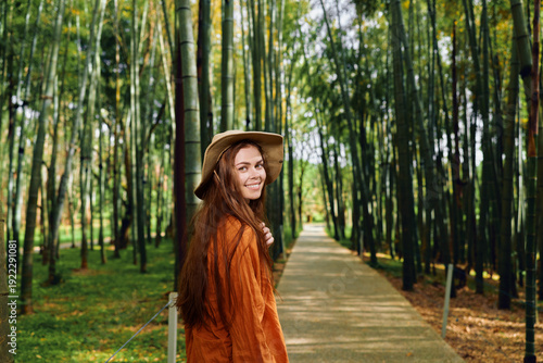 Εκτύπωση καμβά Woman with a warm smile wearing hat and coat walks along a bamboo-lined path and walkway through a forest, portrait of a traveler enjoying nature, green grove and calm outdoor mood