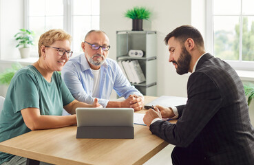 Senior couple signs documents with financial advisor at a desk, using tablet to check contract details as the advisor explains terms before signing. Seniors meeting with agent about insurance. 