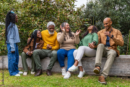 African American family in knit sweaters laughing, sitting on wooden log bench on lawn holding cup