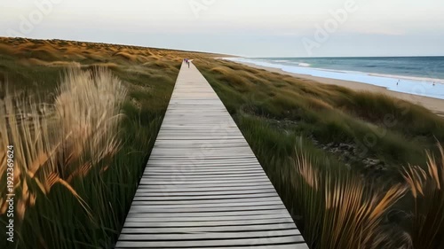 Scenic wooden boardwalk stretches along sandy beach with tall grass on both sides, leading to the ocean under a clear sky at dusk