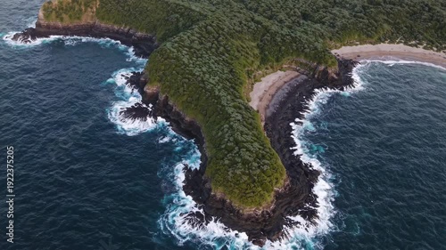 Aerial view of a lush green peninsula extending into the ocean, showcasing rocky shorelines and gentle waves lapping against the sandy beaches in a coastal landscape