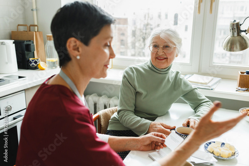 Photography Selective focus on old smiling woman sitting at kitchen table near window and lo