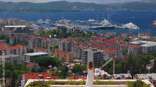 A woman in a white dress stands on a rooftop garden in Montenegro, facing a marina with superyachts and a tall masted vessel, boats moving across a deep bay.