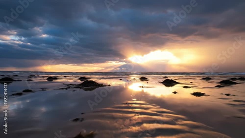 Coastal landscape at sunset with dramatic clouds and sunlight reflecting on wet sand, seaweed scattered across the shore in a serene beach setting