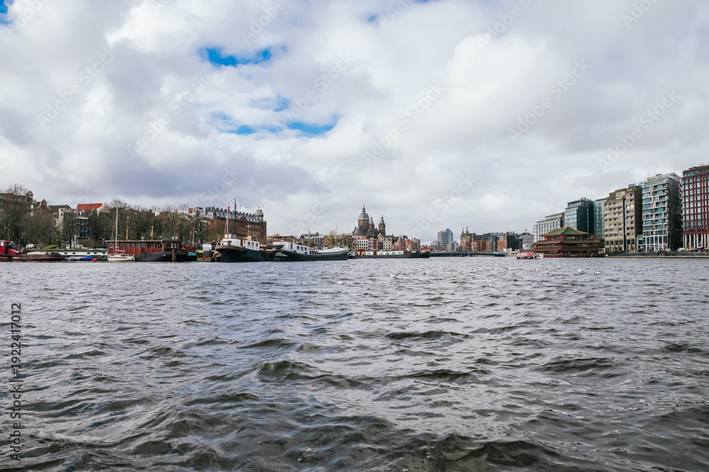Naklejka premium Cityscape of amsterdam showing the amstel river, historic architecture along its banks, modern buildings, and various boats on a cloudy day, creating a panoramic view of urban life and travel