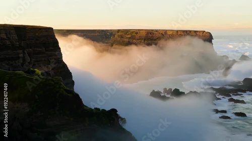 Coastal cliffs with rolling waves and mist rising from the ocean create a dramatic landscape at sunrise, showcasing nature's beauty and tranquility in a serene coastal setting