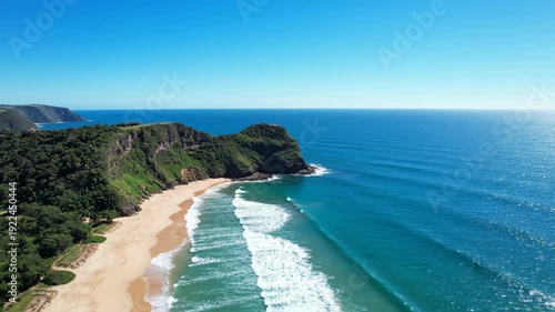 Aerial view of a scenic beach with gentle waves lapping against the shore, surrounded by lush green cliffs and a clear blue sky over a tranquil ocean
