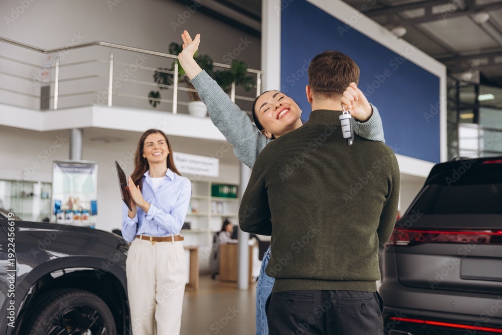 Obraz premium Happy couple hugging after buying new car at dealership, woman holding car key and saleswoman smiling