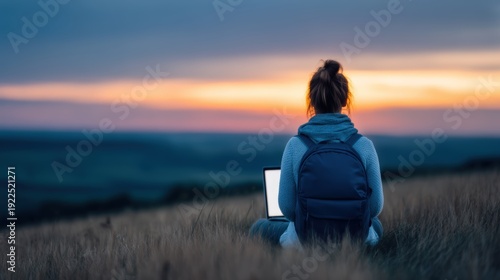 Summer for digital nomads, A person with a backpack sits in a field at sunset, using a laptop and enjoying the peaceful natural landscape.
