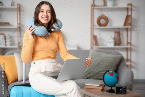 Female traveler with neck pillow and laptop sitting on suitcase at home. Itin...