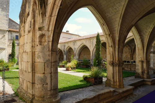 View of the Gothic cloister of Saint-Pierre Cathedral in Saintes, France. The stone arches frame an inner courtyard garden with green grass, plants, and cypress trees under a bright sky