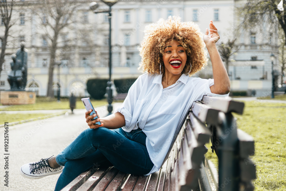 Naklejka premium A joyful young African American woman with a blonde afro expresses excitement and laughter while sitting on a wooden bench in a public park, holding her mobile phone.