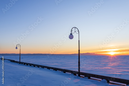 Beautiful winter sunset over the Laurentian mountains and partly frozen St. Lawrence river, with graceful lampposts on village pier in the foreground, Kamouraska, Québec, Canada