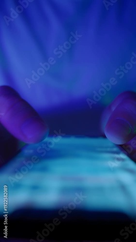 Macro view of man fingers move across a glowing touchscreen, scrolling through social media feed in the dark. Late-night smartphone use or screen dependence. Vertical video