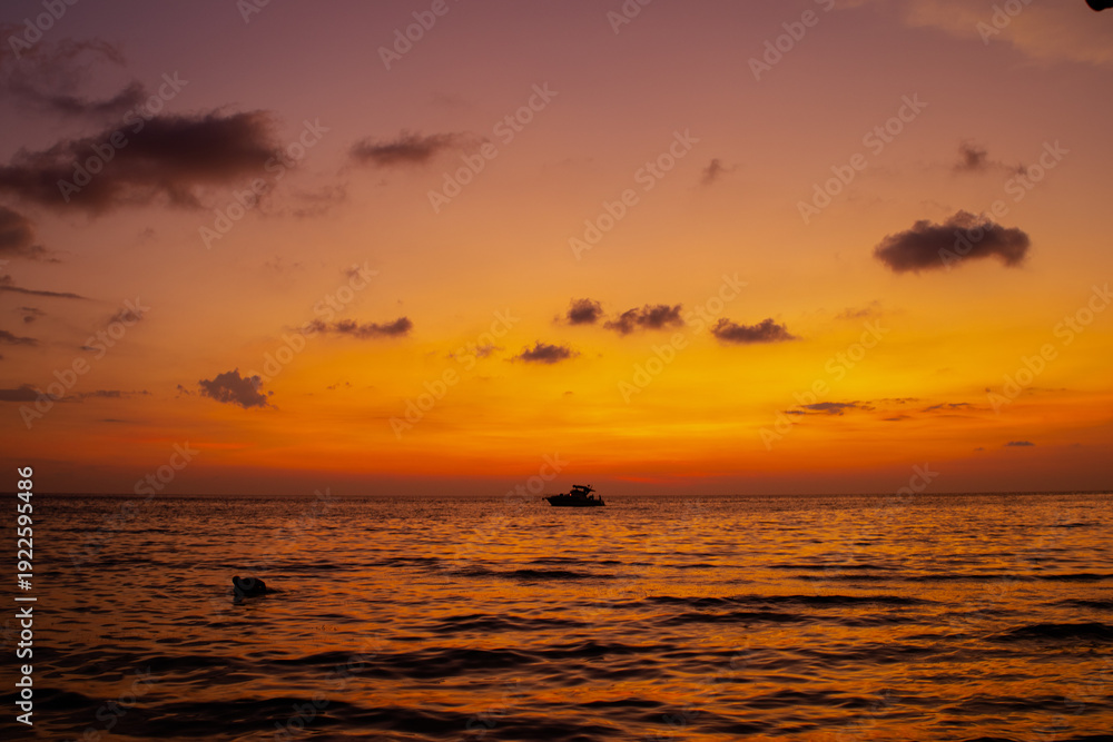 Obraz premium Golden sunset over calm sea with small boat silhouette on horizon. Warm orange sky and sea reflections creating peaceful travel landscape and serene evening atmosphere. Thailand vacation.
