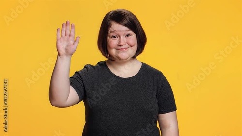 Friendly woman down syndrome waving hello with a happy expression on a vibrant yellow background, showcasing a welcoming gesture.