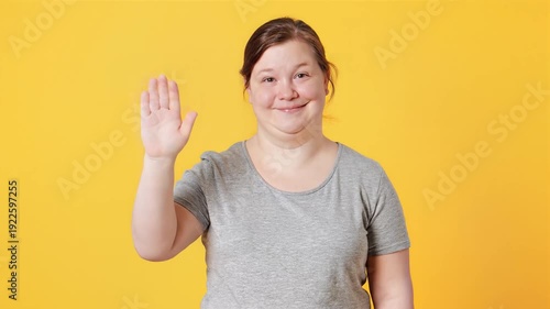 Smiling young woman with Down syndrome waving hand, looking at camera, isolated on yellow background.