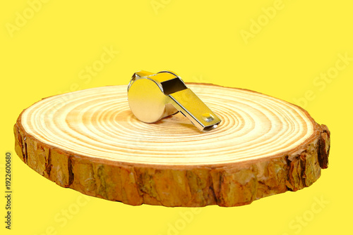 Photos A silver metal referee sports coaching whistle resting upon a natural wooden tree trunk slice podium against yellow background, highlighting themes of authority, guidance, and sound activation