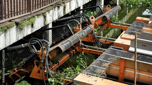 Картината върху платно An orange industrial aquatic weed harvester operating in a canal under a bridge