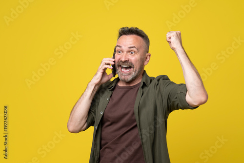 Attractive handsome 50s man over isolated yellow background have mobile conversation over smartphone in victory position. casual man hearing excelent news over phone and celebrating success with fire