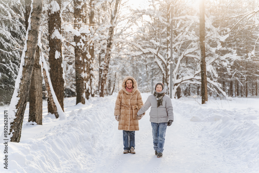 Fototapeta premium Walking mother with teen son on snowy forest road, tall trees, calm winter landscape, family time, bonding, childhood memory, outdoor activity, healthy lifestyle, weekend leisure, holiday mood.