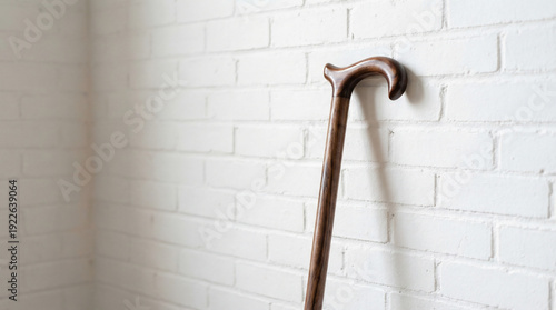 Wooden cane with curved handle leaning against white brick wall with soft diagonal shadow and subtle texture.
