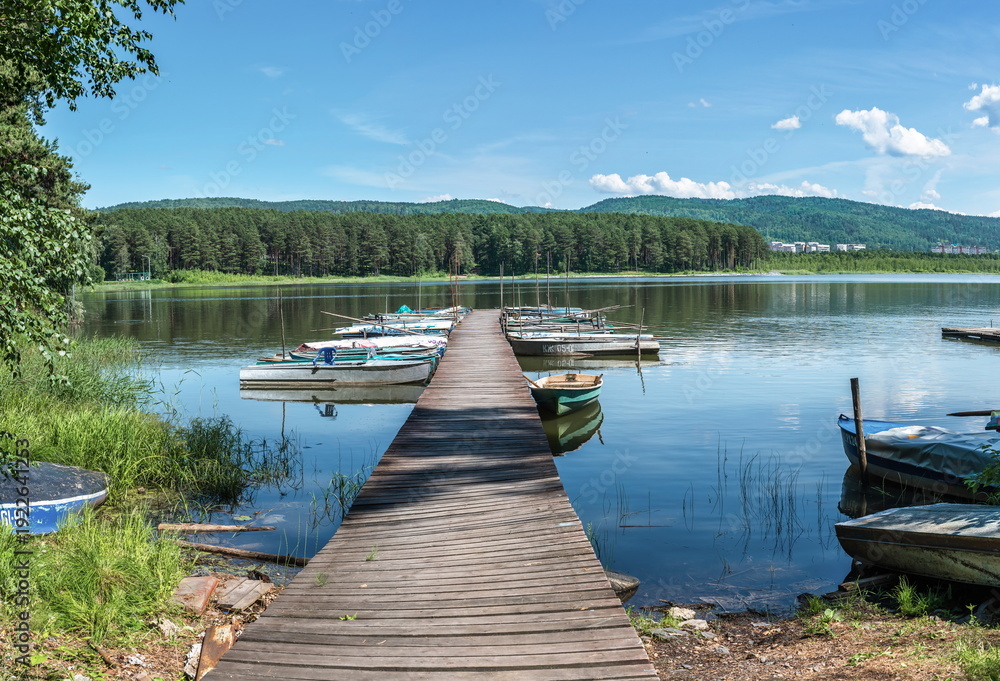 Fototapeta premium A wooden pier extending into the lake, with moored boats. There is water, greenery and for