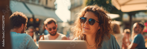 A young woman wearing sunglasses smiles while working on a laptop outdoors at a sunny cafe with people chatting around her.
