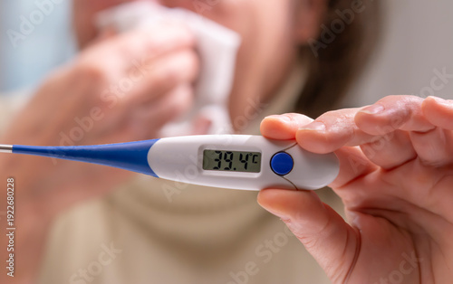A man wiping his face with a napkin displays a high temperature of 39.4 degrees Celsius on an electronic thermometer.